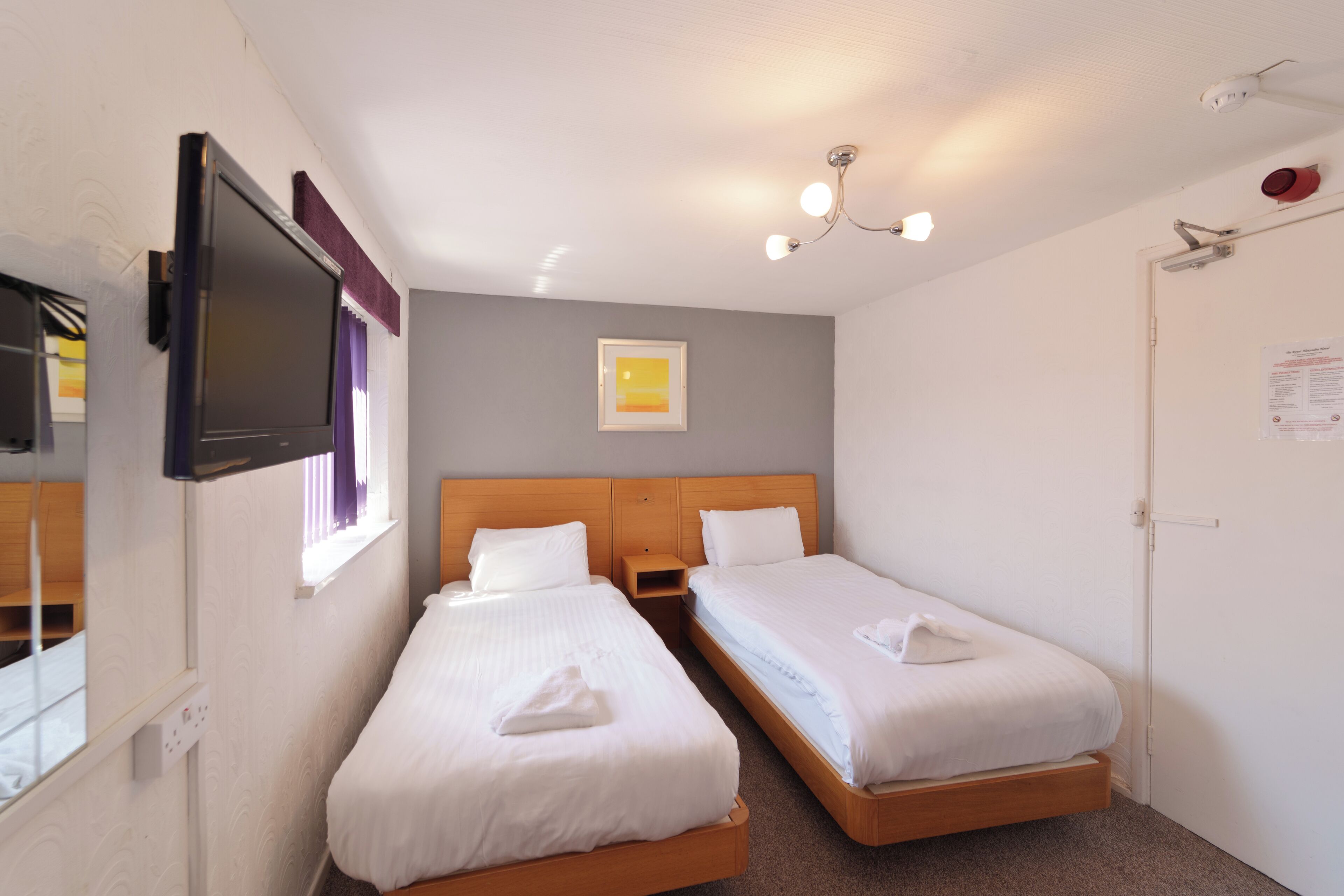Habitación de hotel con dos camas individuales, un televisor montado en la pared, una lámpara en el techo y una puerta con un cartel.