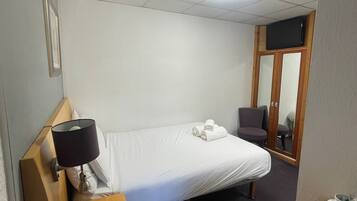 Hotelkamer met een enkel bed, nachtkastje met lamp, stoel, bureau met beker en afstandsbediening, en een deur naar de badkamer.