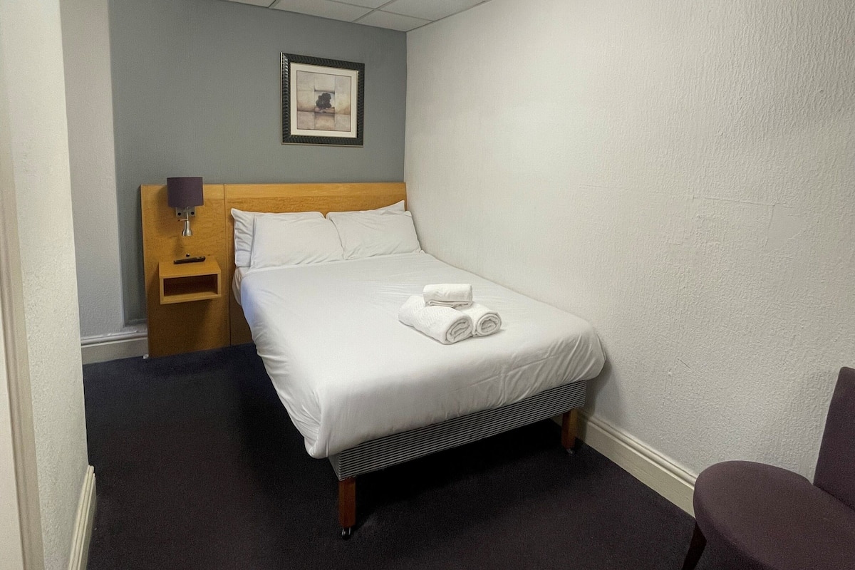 Habitación de hotel con una cama individual, una mesita de noche, una silla, un cuadro en la pared y una lámpara en el techo.
