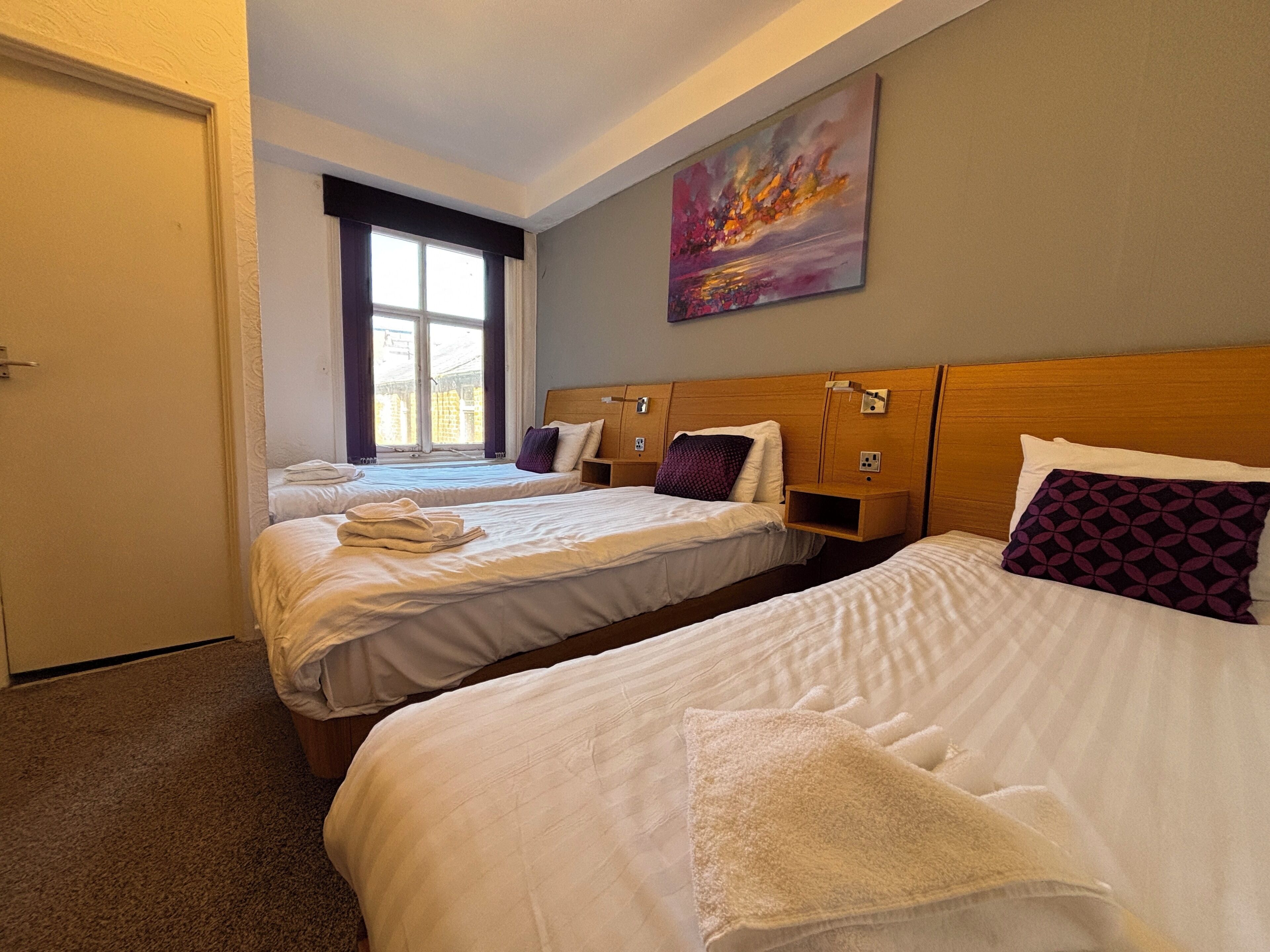 Ein Hotelzimmer mit zwei Betten, einem Vorhangfenster, einem Gemälde an der Wand und einer Tür.