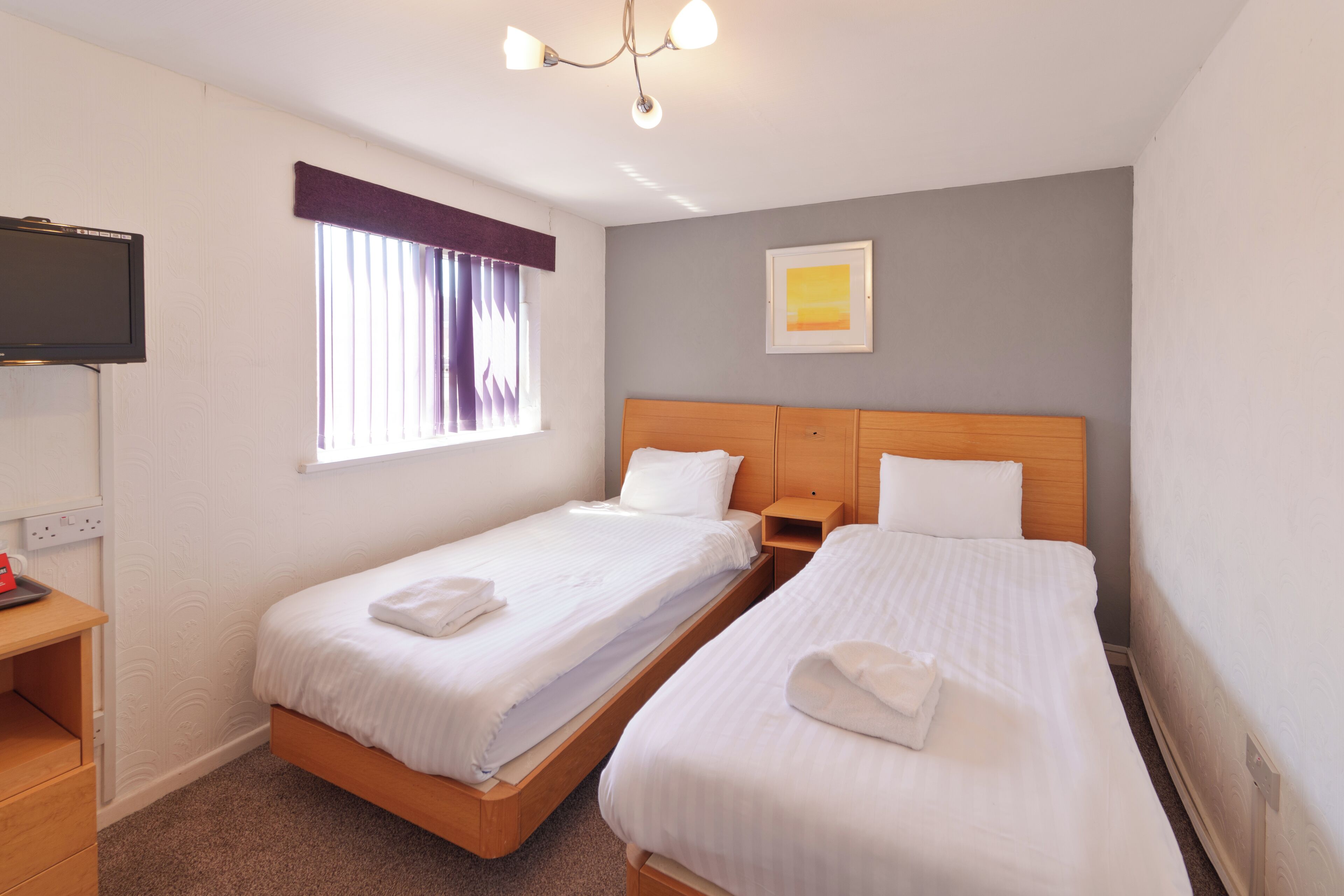 A hotel room with two single beds, a TV, a window with curtains, a painting on the wall, and a wooden nightstand.
