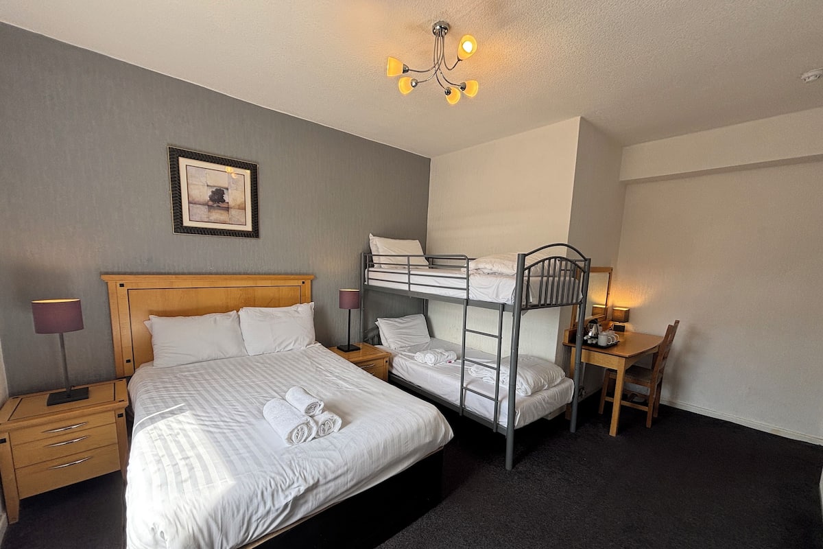 Habitación de hotel con una cama individual, una litera, un escritorio con silla, una mesita de noche, una lámpara y un cuadro en la pared.
