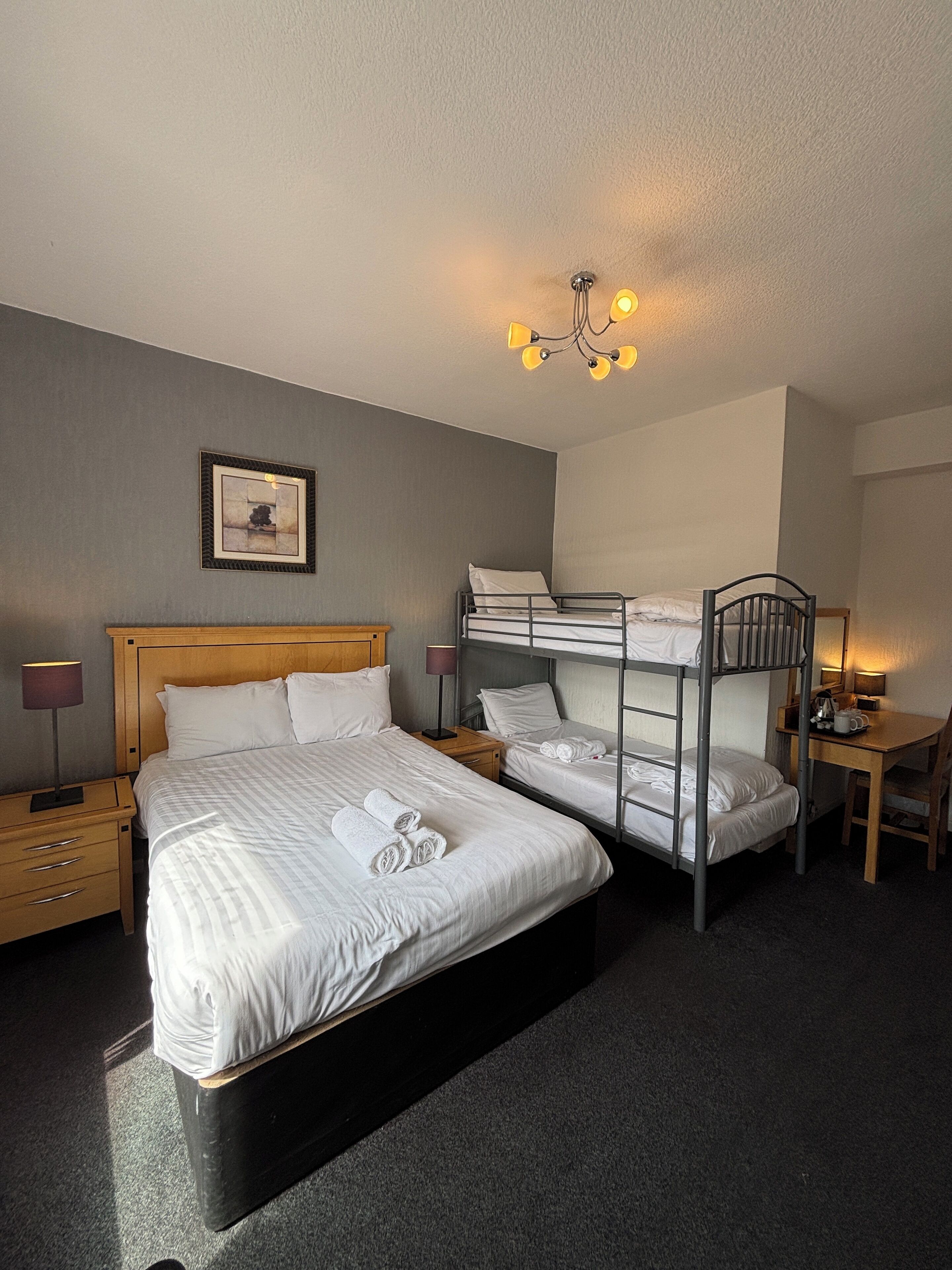 Quarto de hotel com uma cama de solteiro, uma cama beliche, uma escrivaninha com cadeira, mesa de cabeceira com abajur e um quadro na parede.