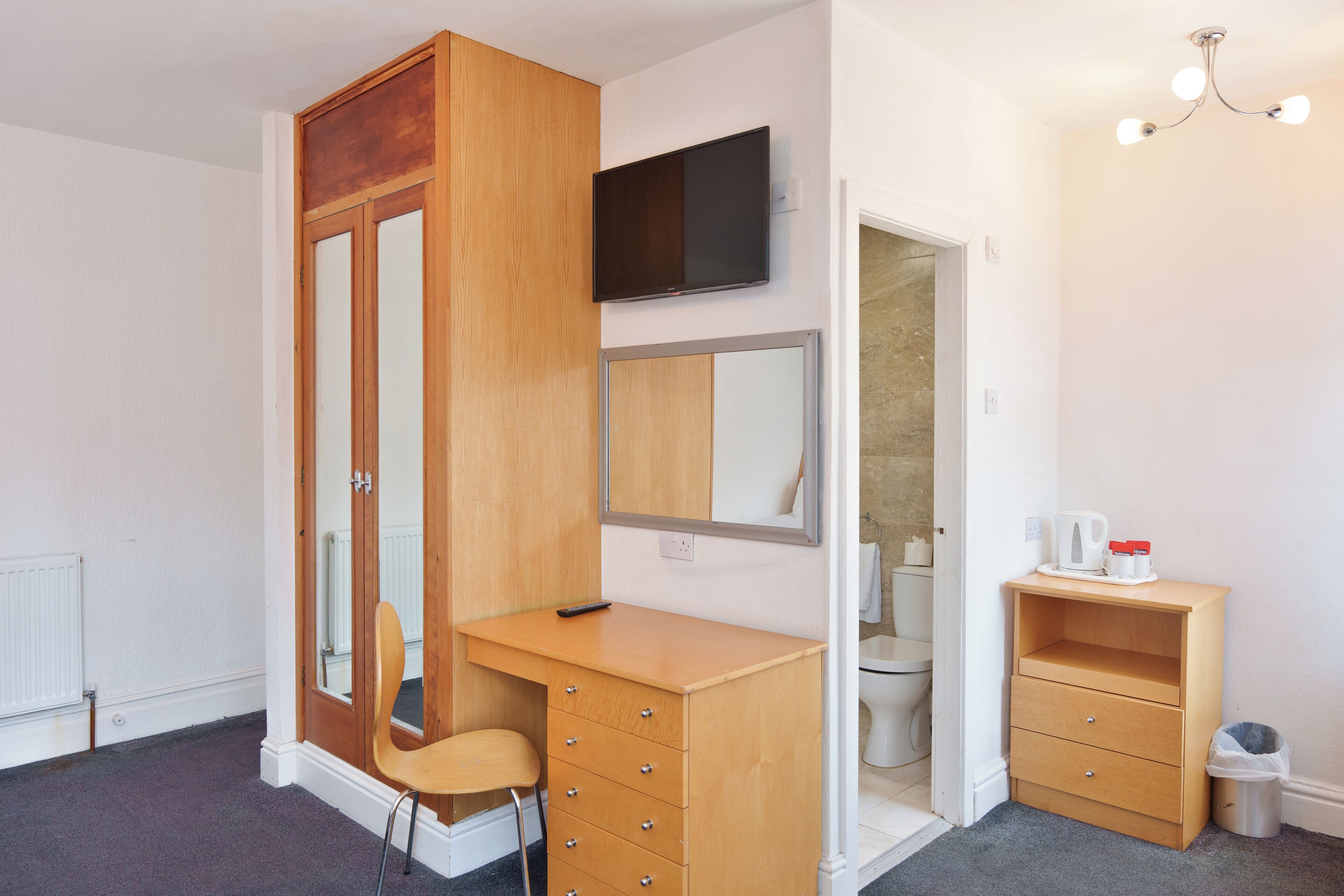 A bedroom with a wooden wardrobe, a desk with drawers, a chair, a television mounted on the wall, and a small shelf with a kettle and cup.