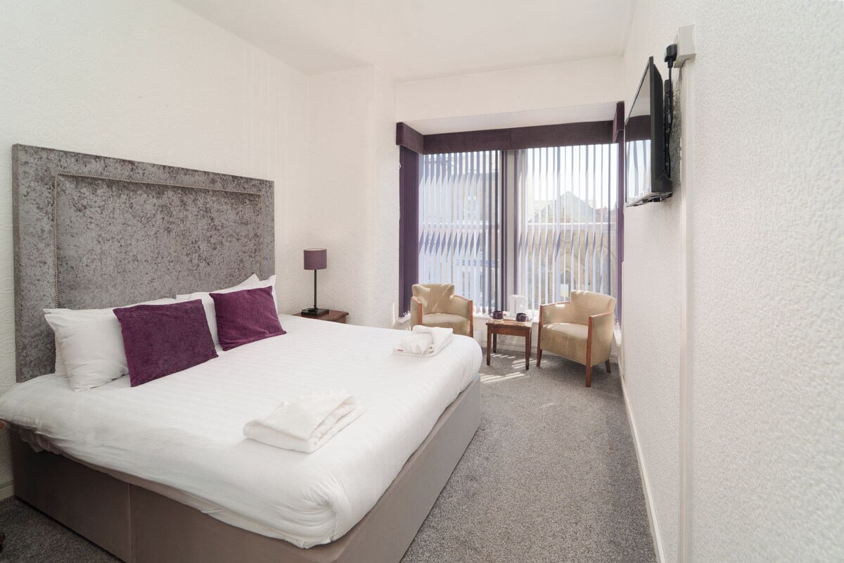 A hotel room with a large bed, a TV mounted on the wall, a chair, and a small table.