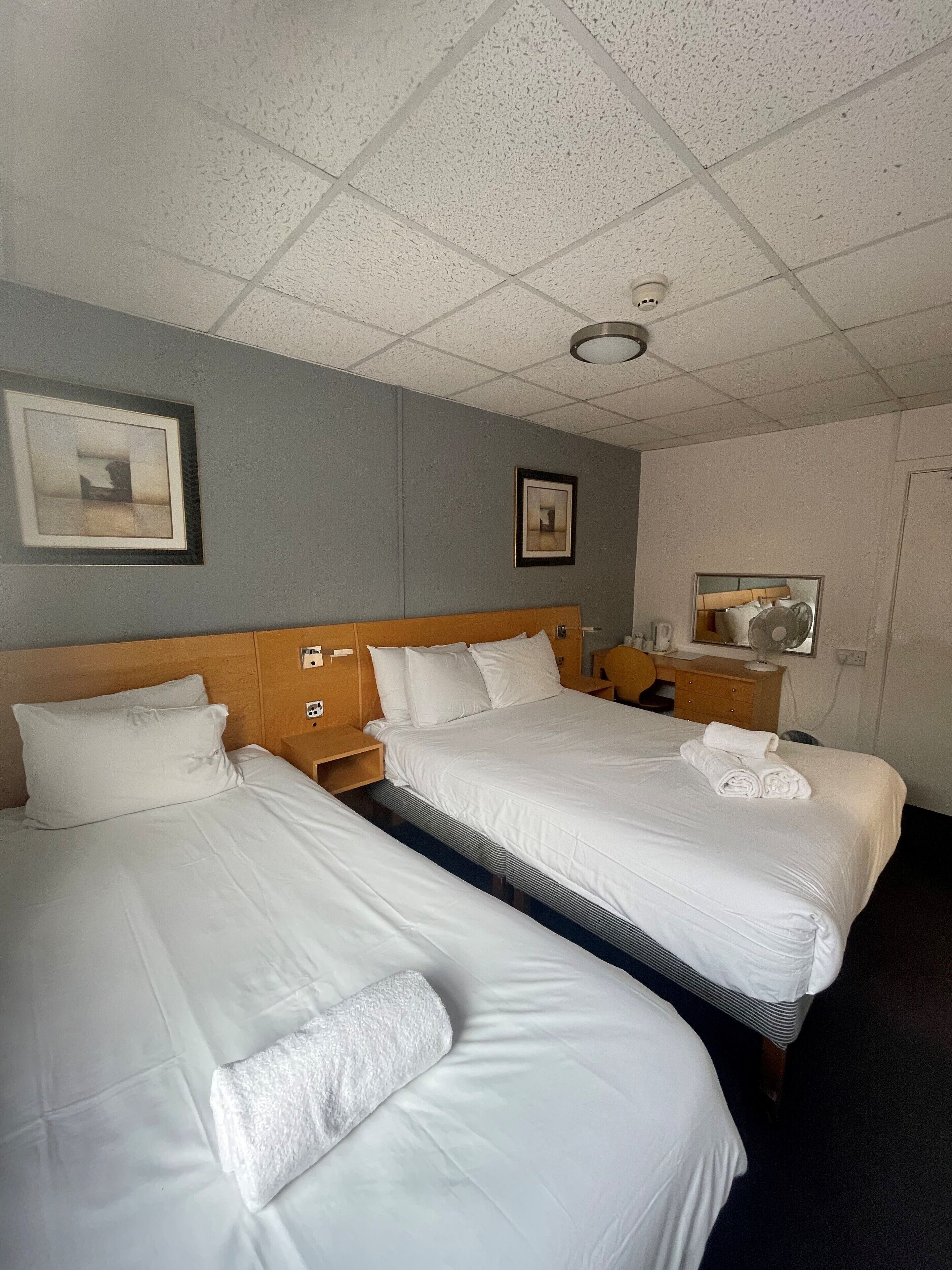 A hotel room with two beds, a wooden headboard, a bedside table with a lamp, a framed picture on the wall, and a mirror on the opposite wall.