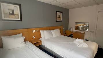 A hotel room with two beds, a wooden headboard, a bedside table with a lamp, a framed picture on the wall, and a mirror on the opposite wall.
