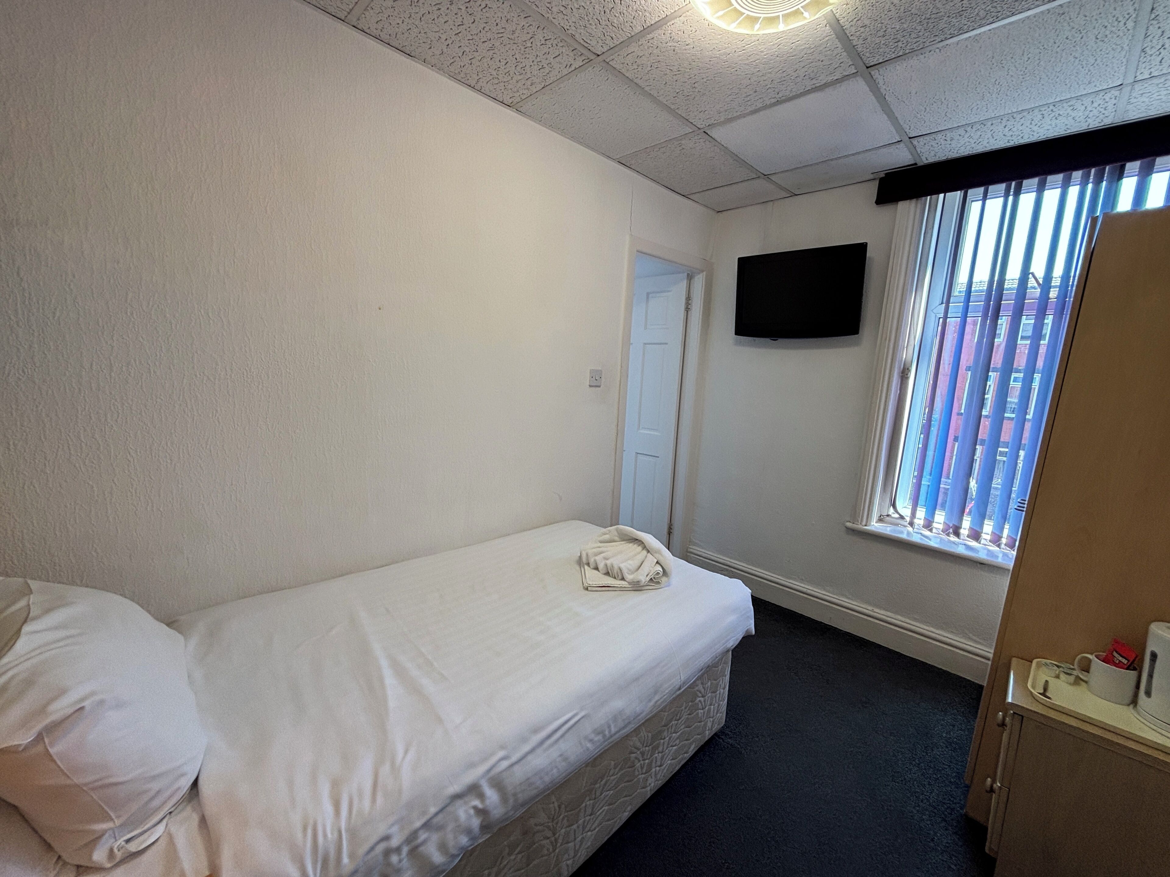 A small, clean room with a single bed, a flat-screen TV on the wall, and a window with blinds.