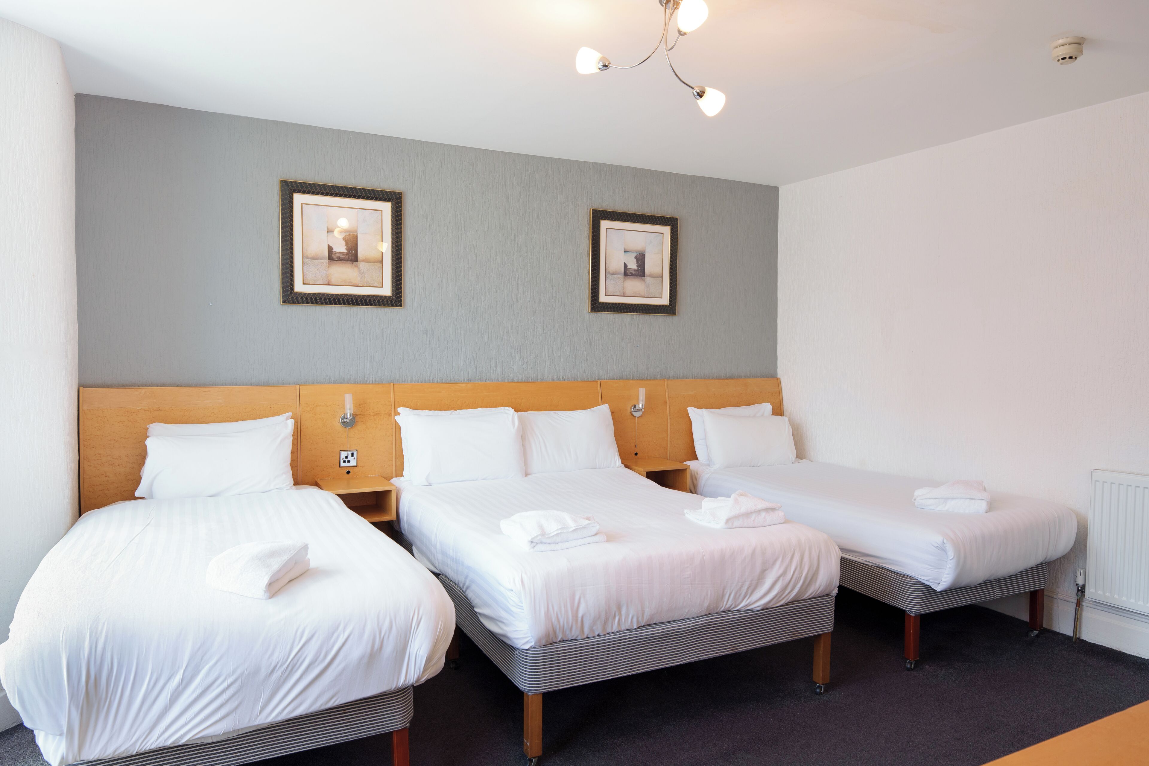 Une chambre d’hôtel avec trois lits simples, une tête de lit en bois et deux tableaux encadrés au mur.