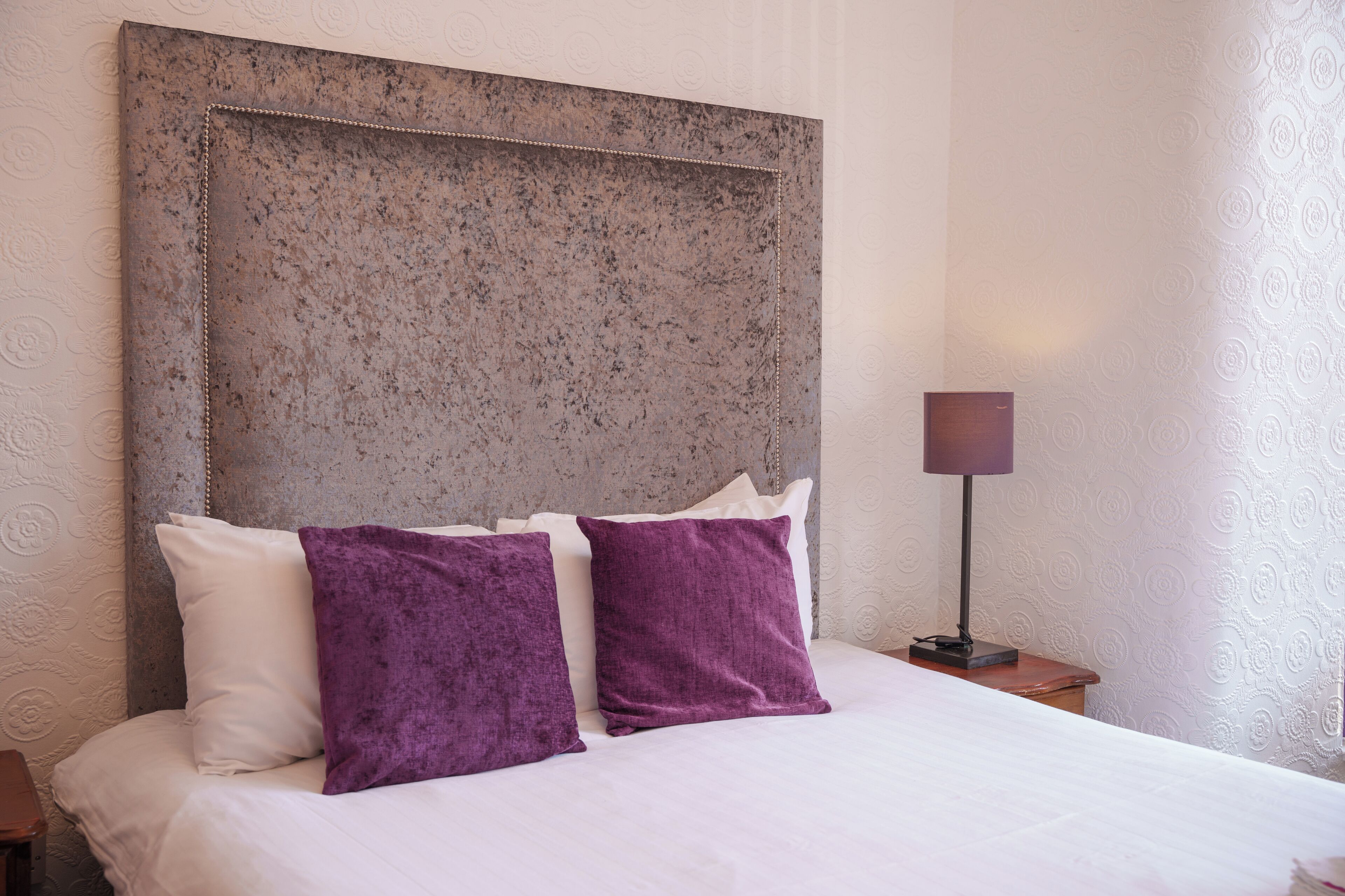 A neatly made bed with white and purple pillows, a textured headboard, and a bedside lamp on a nightstand.