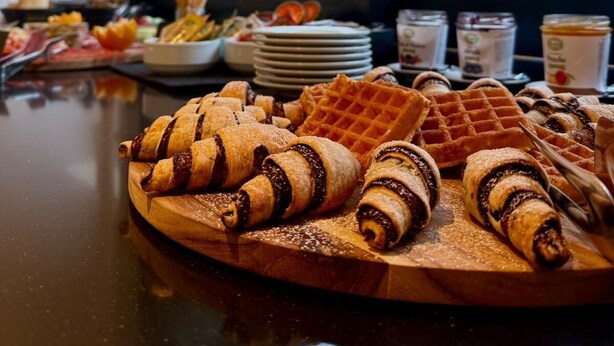 A breakfast buffet with pastries and waffles on a wooden board, with a window view of trees. - B&B Arena Prestige Rooms (Pula)