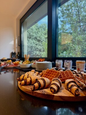 Un buffet de petit-déjeuner avec des viennoiseries et des gaufres, présenté sur une planche en bois, avec vue sur les arbres par la fenêtre.