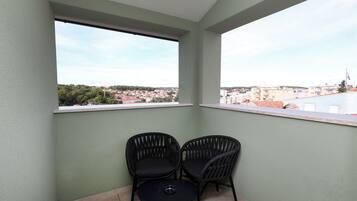 A balcony with two wicker chairs and a small table, overlooking a cityscape with buildings and greenery.
