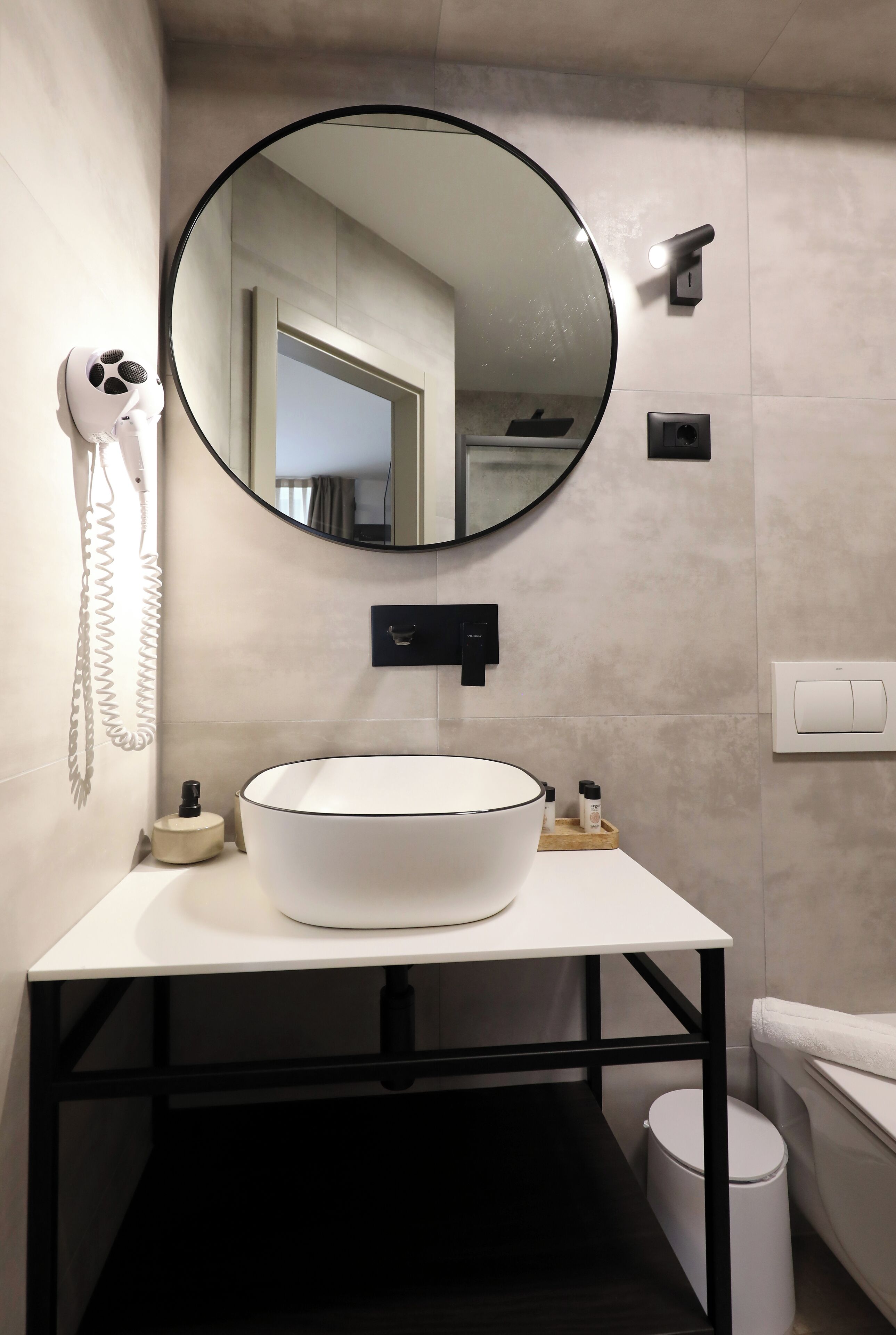 A modern bathroom with a round mirror, a white sink, a hairdryer, and a toilet.