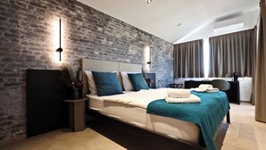 A modern bedroom with a brick wall, a large bed with teal bedding, a bedside table with a vase, and a desk with a chair. - B&B Arena Prestige Rooms (Pula)