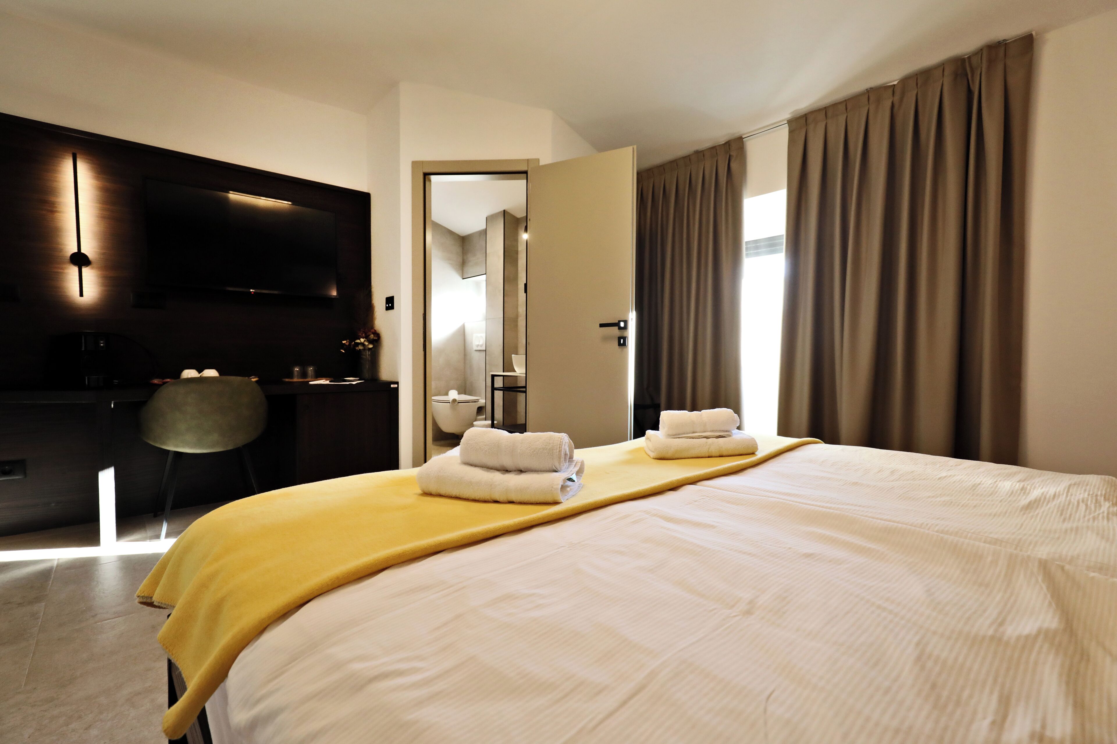 Une chambre d’hôtel avec un grand lit, un bureau avec une chaise, un téléviseur à écran plat fixé au mur et une salle de bain visible à travers une porte ouverte.