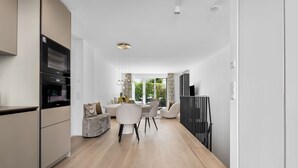 A modern kitchen with built-in appliances, a dining area with a round table and chairs, and a view of a garden through large windows. - Luxury duplex apartment with terrace (Sylt)