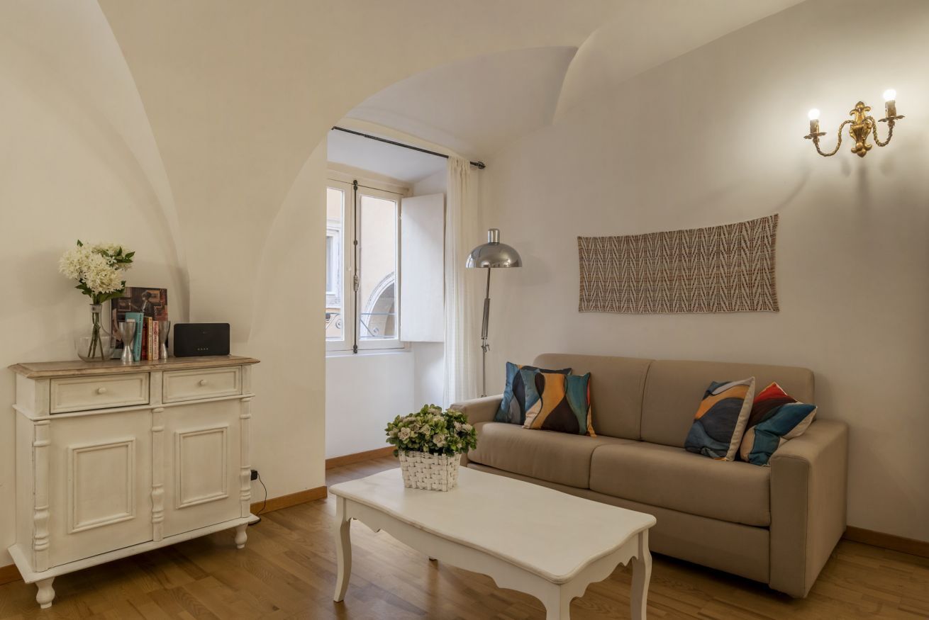 Una sala de estar con un sofá color beige, una mesa de centro blanca y un mueble blanco. Hay una lámpara, un aplique de pared y un tapiz con diseño.