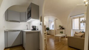 A compact kitchen with a central island, a dining area with a white table, and a living space with a sofa and a window.