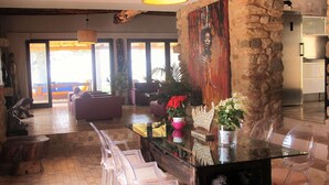 A spacious living area with a long glass dining table, clear plastic chairs, a stone accent wall, and a large window providing natural light. - My Rental Homes - Villa Sun with sea view, pool and large garden (Sant Antoni De Portmany)