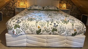 A bedroom with a stone wall, a bed with a floral bedspread, two bedside lamps, and a window. - Gîte "la Cazelle (Saint-Chels)