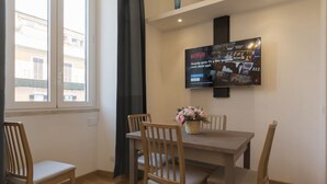A dining area with a table and four chairs, a TV mounted on the wall displaying Netflix content, and a window with a view of buildings outside. - CS Termini Nazionale Apt n. 4 (Roma)
