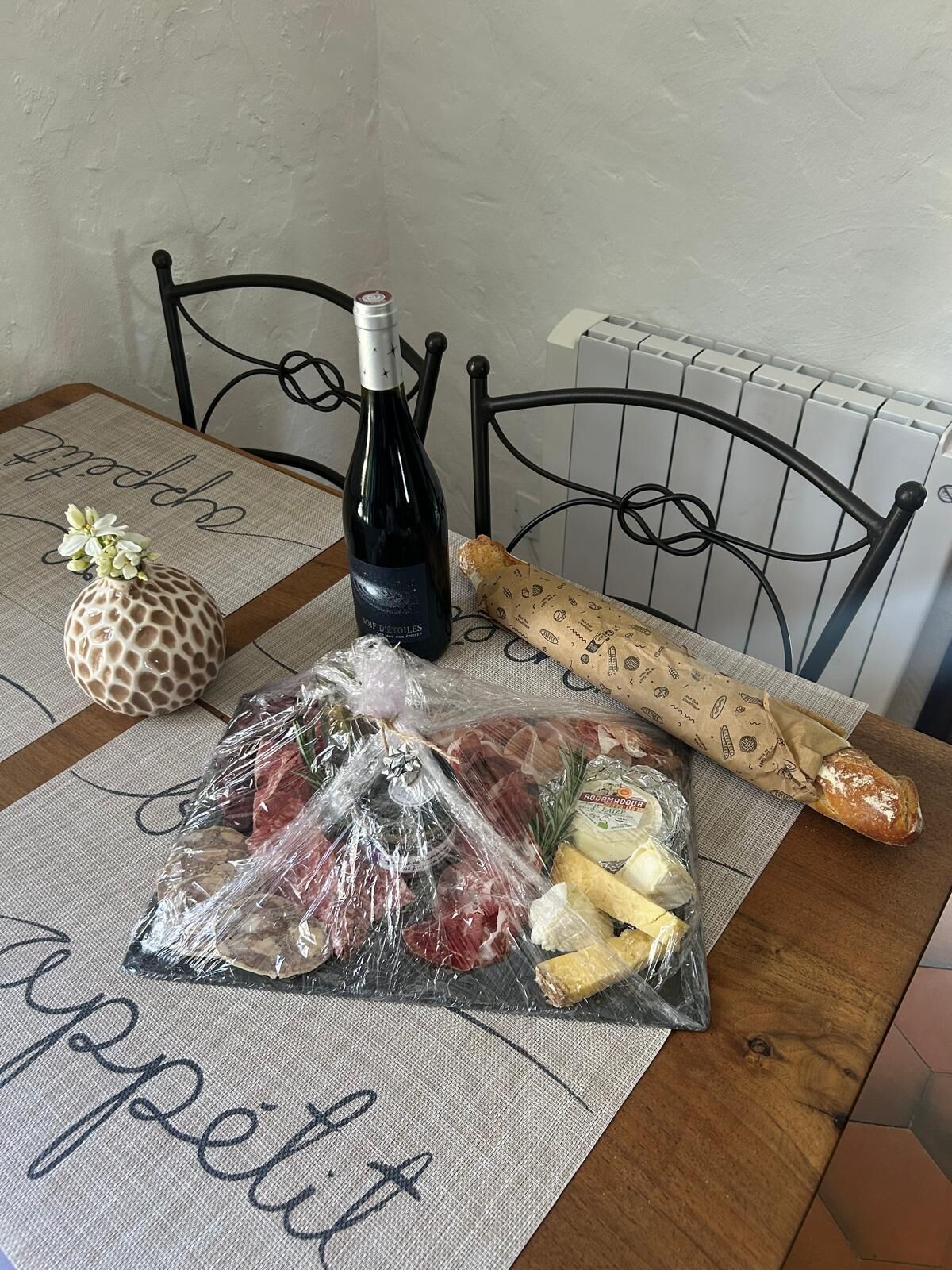 A table with a bottle of wine, a baguette, a wrapped ham, cheese, and a decorative item.