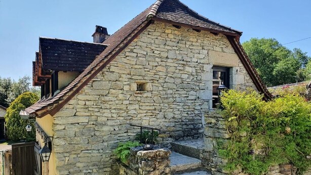 A stone building with a sloped roof, a small staircase leading to a door, and a garden with flowering plants. - Gîte "Le Causse (Saint-Chels)