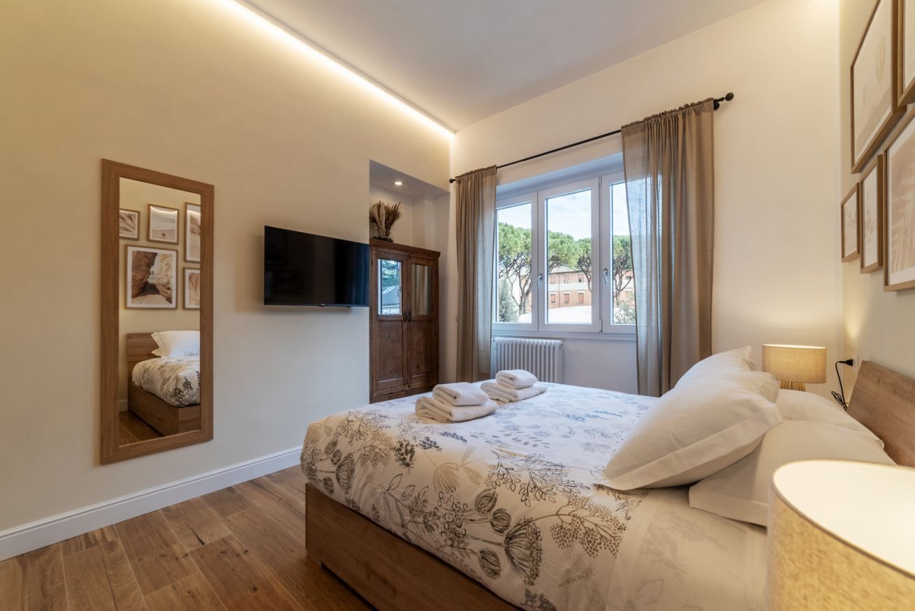 Una camera da letto con un letto, una TV a schermo piatto, uno specchio, una finestra con tende e un termosifone.