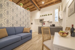 Un salon avec un canapé bleu, une table à manger en bois sur laquelle se trouve un panier de fruits, et une cuisine en arrière-plan.