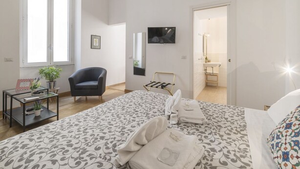 A bedroom with a bed, a chair, a TV, a mirror, and a bathroom visible through an open door.