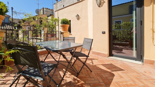 An outdoor patio with a glass table and chairs, surrounded by potted plants and a brick floor. - CS Stylish Loft with Terrace (Roma)