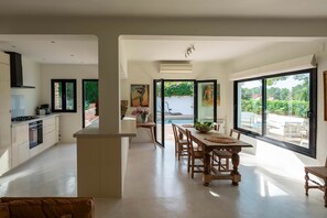 A modern kitchen with a central island, a dining area with wooden chairs, and large windows offering a view of the outdoors. - Can Blanca with pool - My Rental Homes (Sant Antoni De Portmany)