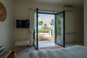 A bedroom with a bed, a TV, a chair, and an open door leading to a balcony with a view of palm trees and a garden. - Can Blanca with pool - My Rental Homes (Sant Antoni De Portmany)