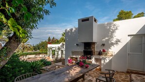 Eine Terrasse im Freien mit Holztisch und -stühlen, einem eingebauten Grill und einer weißen Wand mit einem Fenster.
