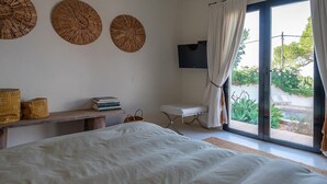 Ein Schlafzimmer mit Bett, einem an der Wand befestigten Fernseher, einem Fenster mit Vorhängen und einem kleinen Tisch mit Büchern und Körben.