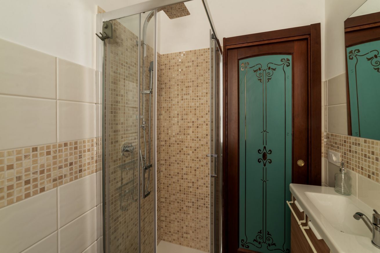 A bathroom with a glass shower enclosure, a wooden door with decorative metalwork, and a white sink with a mirror.
