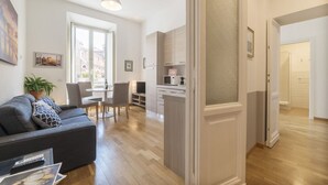 A modern living room with a sofa, a dining table, and chairs. There is a kitchen area with cabinets and a microwave. The room has wooden flooring and a large window. - CS Central Station Termini / Monti Apartment (Roma)
