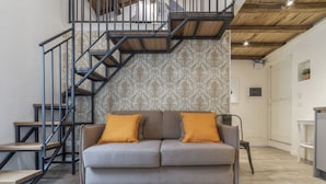 Een moderne woonkamer met een grijze bank, twee oranje kussens, een houten trap en behang met een patroon.
