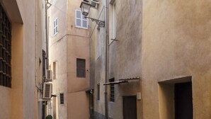 A narrow street with buildings on either side, featuring air conditioning units, windows, and a small plant.