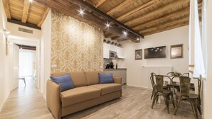 A living room with a sofa, dining table, and chairs. There is a kitchen area with a microwave and a coffee machine. The room has wooden beams on the ceiling and patterned wallpaper.