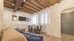 A cozy living room with a sofa, a dining table with chairs, a TV mounted on the wall, and a wooden ceiling.