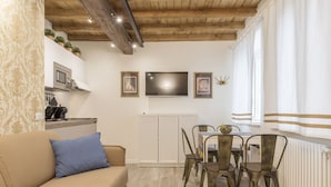 A cozy living room with a beige sofa, a dining table with four chairs, a flat-screen TV mounted on the wall, and a kitchen area with a microwave and cabinets.