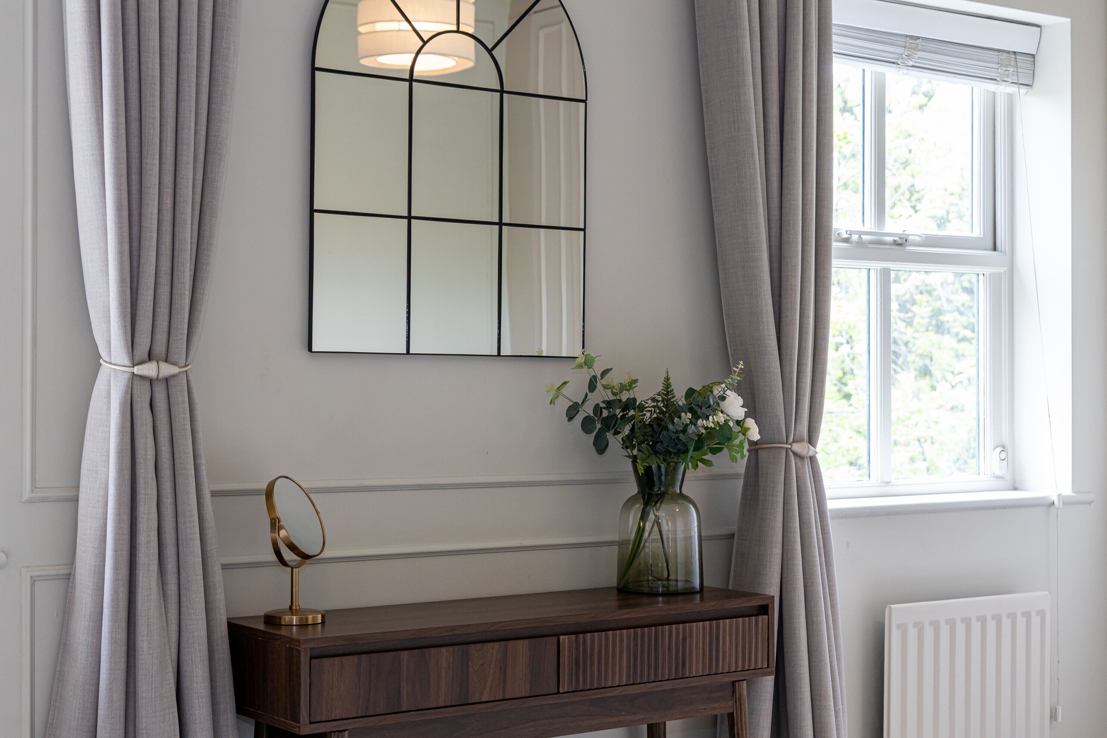 A room with a wooden console table, a mirror, a vase with flowers, and a window with curtains.