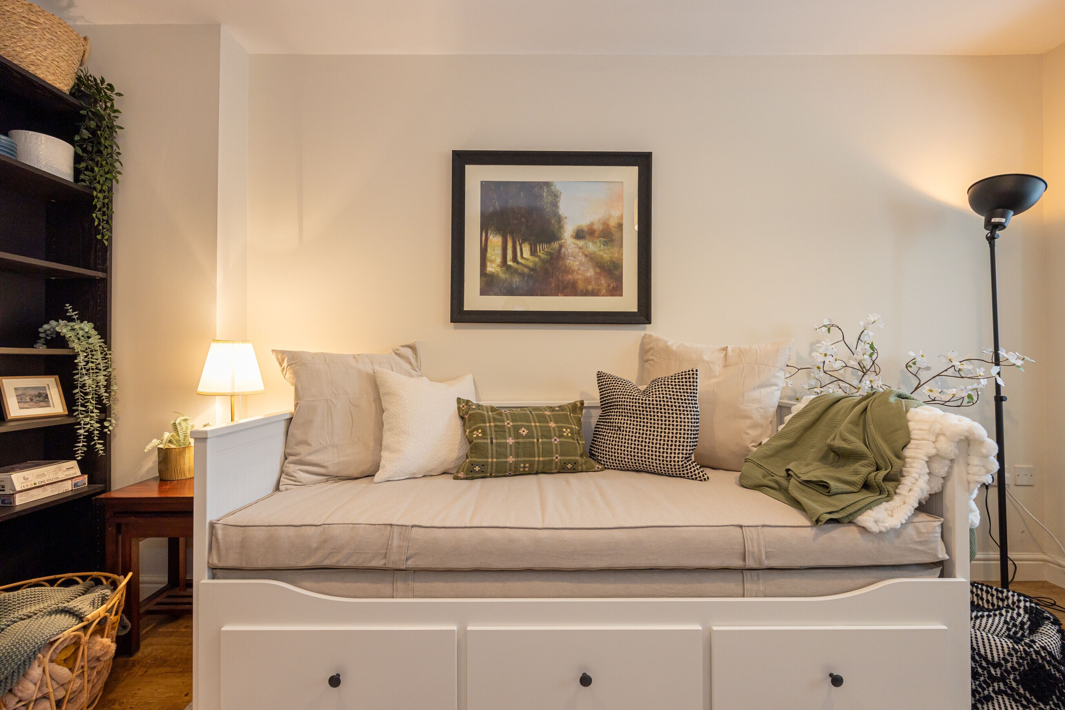 A cozy living room with a sofa, a framed painting, a lamp, and a bookshelf.