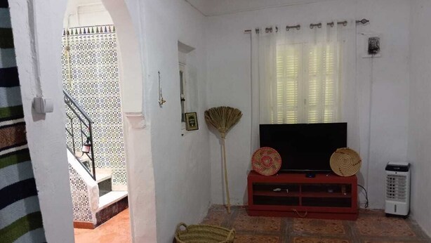 A living room with a TV on a red stand, a standing fan, and a woven basket on the floor. - The bird house (Tlemcen)