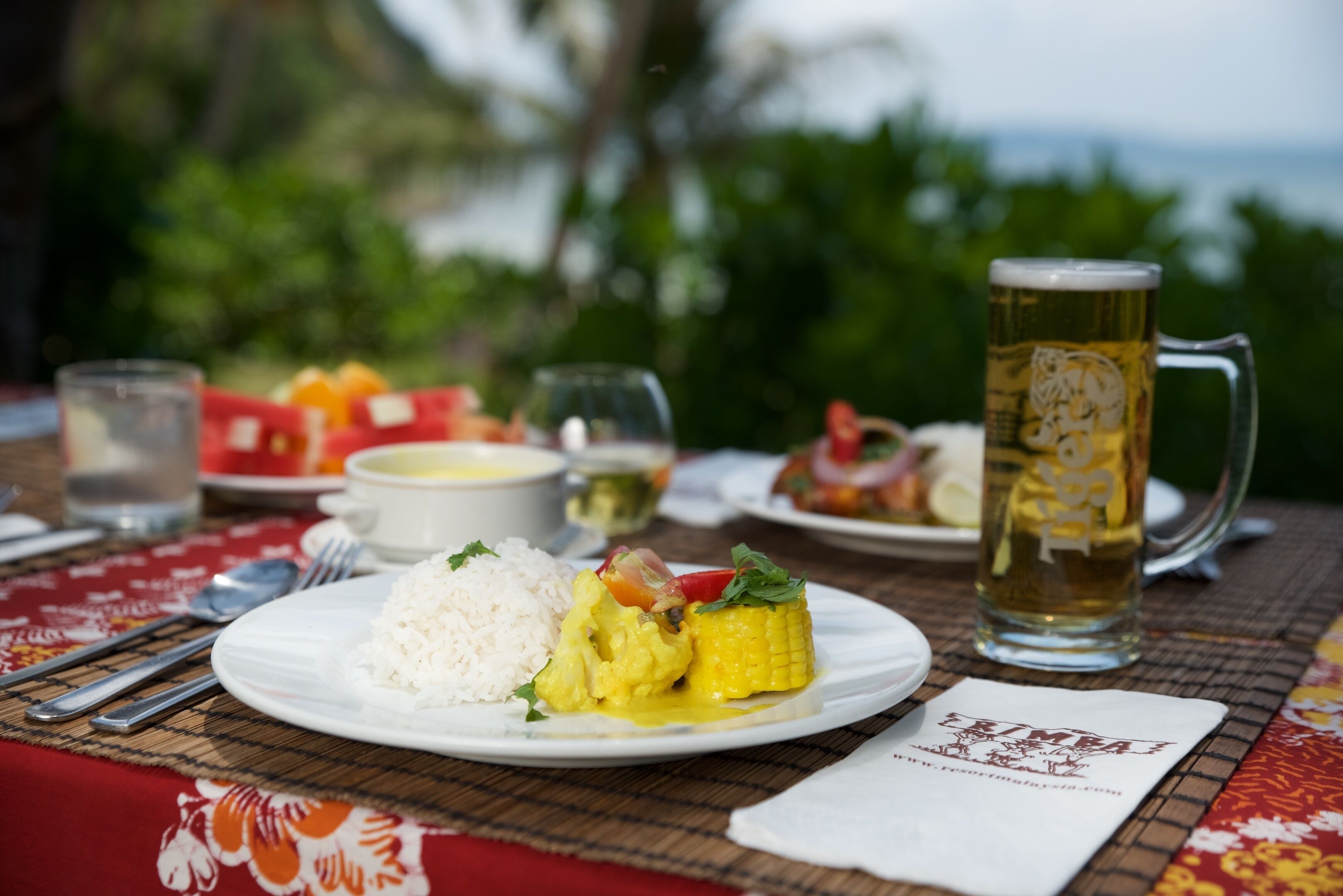 Un plat contenant du riz, du maïs et des légumes, une chope de bière et un verre d’eau, le tout posé sur une table recouverte d’une nappe rouge.