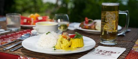 A meal with rice, corn, and vegetables on a plate, a beer mug, and a glass of water on a table with a red tablecloth.