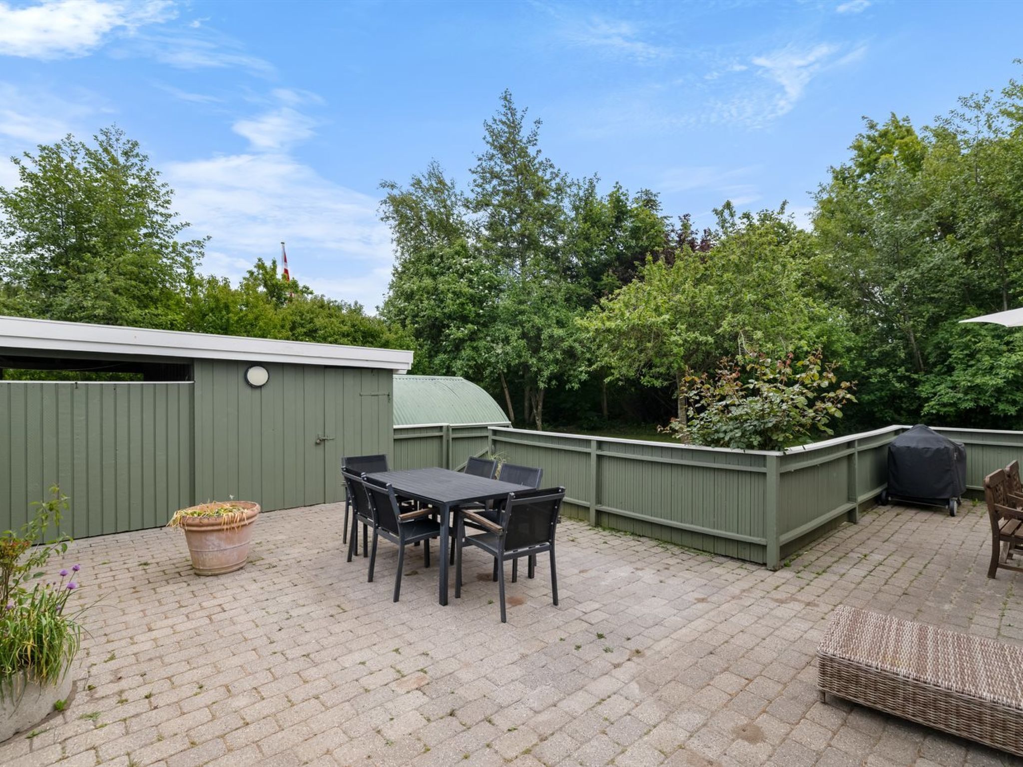 An outdoor patio area with a table and chairs, a covered grill, and a green fence.