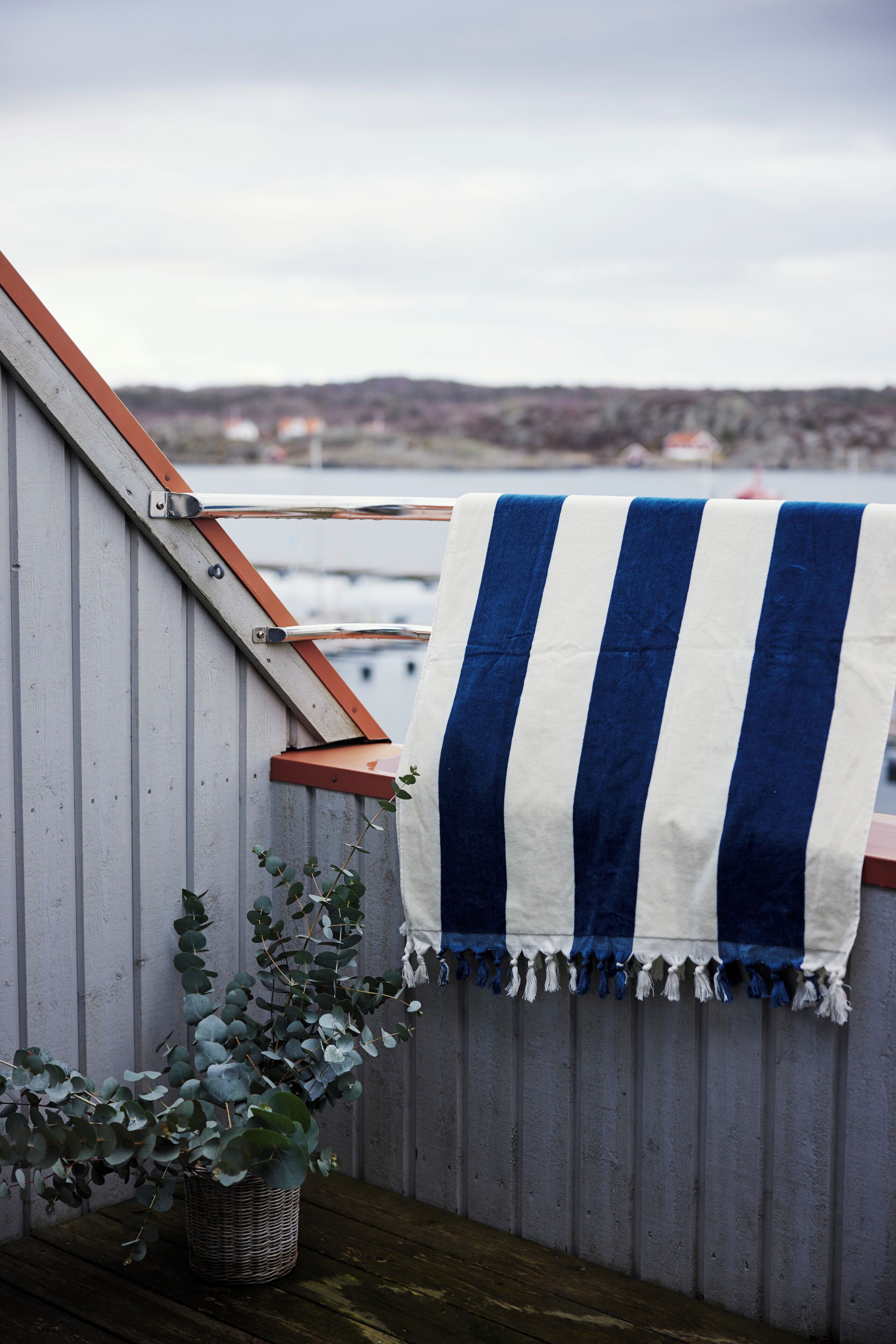 A balcony with a striped towel, a wicker basket with plants, and a view of boats and hills in the background.